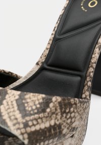 Közelkép egy kígyóbőr mintájú magassarkú szandálról, párnázott fekete talpbéléssel és nyitott orrkialakítással.