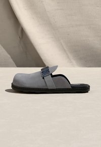 Γκρι παπούτσι τύπου slip-on με μαύρη σόλα και ρυθμιζόμενο λουράκι με κομμένα γράμματα, τοποθετημένο πάνω σε ουδέτερο φόντο από ύφασμα και επιφάνεια.