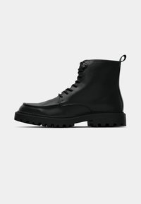 Botte de combat noire en cuir avec lacets, semelle épaisse à crampons et languette de tirage à l’arrière, vue de côté sur un fond uni.