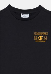 Μαύρο βαμβακερό μπλουζάκι με στρογγυλή λαιμόκοψη, κοντά μανίκια και κίτρινα γραφικά με κείμενο. Το σχέδιο περιλαμβάνει το "CHAMPION 1919" κ αι ένα λογότυπο.