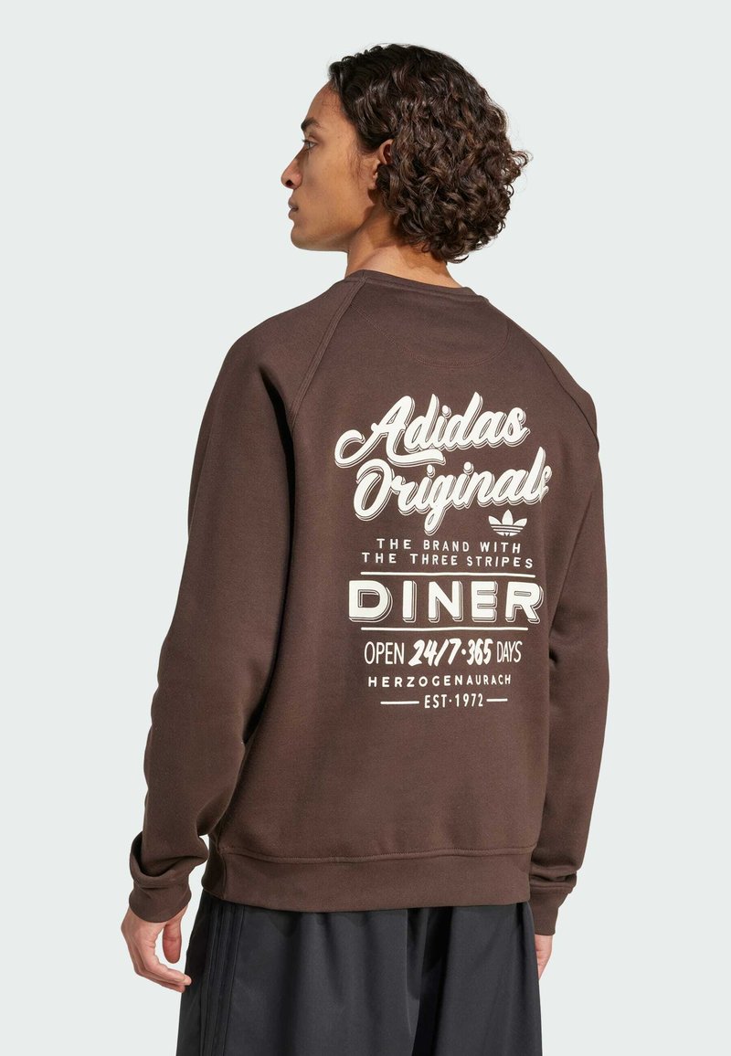 Hnědá mikina s bílým potiskem na zadní straně, včetně textu "Adidas Originals" a detailů z jídelny. Vyrobena z měkkého, strukturovaného materiálu.