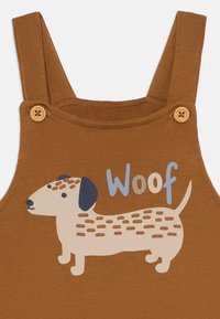 Brune bomullssuspendere med et lekent dachshund-trykk, pyntet med teksten "Woof" i blått, og med trebistropler på stroppene.