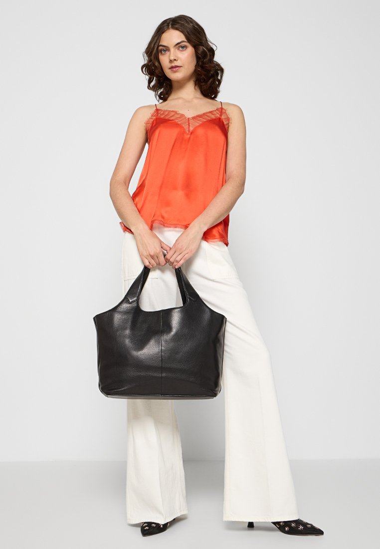 Eine Frau steht und hält eine große schwarze Ledertasche, trägt ein leuchtend orangefarbenes ärmelloses Oberteil, weiße weite Hosen und schwarze spitze Schuhe.