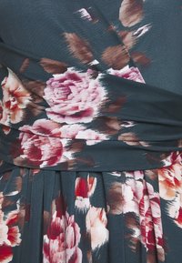 Florales Wickelkleid aus dunkelpetrolfarbenem Stoff mit großen pinken und roten Blumen sowie braunen Akzenten. Der Rock ist mit Falten verziert.