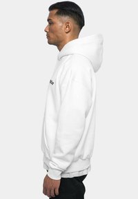 Sweat-shirt à capuche blanc avec poche frontale, poignets et ourlet côtelés. Présente un texte noir sur la poitrine. Texture lisse et design à coupe décontractée.