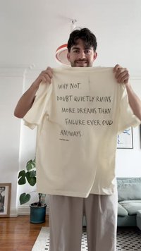 Crème-kleurig, T-shirt met korte mouwen in een relaxte pasvorm, voorzien van zwarte tekst die zegt: "WAAROM NIET. TWIJFELEN VERNIETIGT MEER DROMEN..."