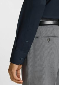 Mörkblå skjorta med lång ärm och knäppna ärmar, kombinerad med grå byxor med struktur och en diskret rutad mönstring samt ett svart läderbälte.