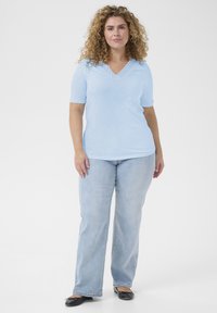 Top corto de color azul claro con escote en forma de concha y detalle de ojales, combinado con jeans azul claro y zapatos planos negros.