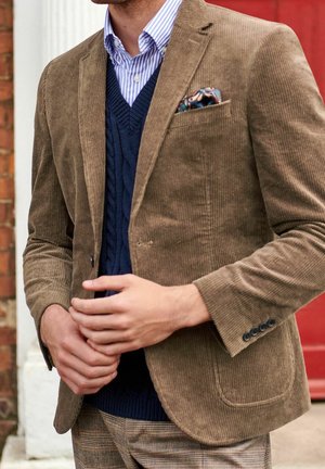 Homme portant un blazer en velours côtelé marron, un pull bleu à torsades, une chemise rayée, un pantalon à carreaux et un mouchoir de poche à motifs, ajustant les boutons du blazer.