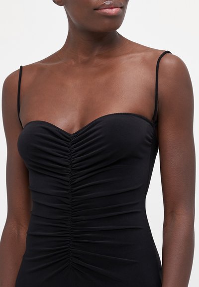 Schwarzes, tailliertes Kleid mit dünnen Trägern, das eine Raffung an Grenze und Taille aufweist, aus glattem, elastischem Stoff gefertigt.