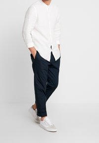 Camicia bianca di lino a maniche lunghe con colletto a fascia, abbinata a pantaloni blu navy e scarpe da ginnastica bianche, che mostrano una vestibilità relax e uno stile casual.