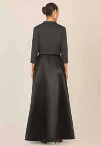Musta satiinimekko, jossa on istuva lyhyt jakku, kolmangin hihat ja pitkä, virtaviivainen hame. Kankaan pinta on sileä ja kiiltävä.