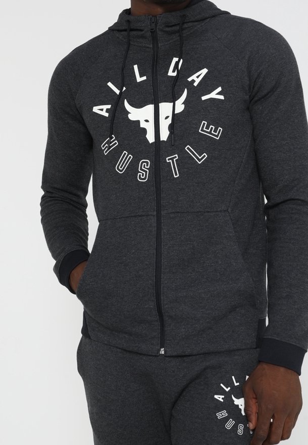 Homme portant un sweat à capuche zippé gris foncé et un pantalon assorti avec un logo de taureau blanc et le texte "All Day Hustle" sur la poitrine et la cuisse.