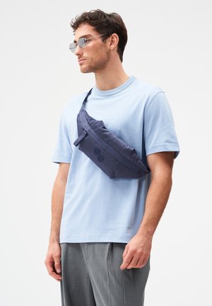Mann mit runden Sonnenbrillen, hellblauem T-Shirt, grauer Nadelstreifenhose und einer navyblauen Umhängetasche über der Schulter.