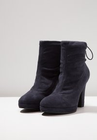 Paire de bottines en daim noir, à talons carrés hauts et bouts ronds, posées sur une surface blanche.