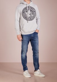 Sudadera gris con un logo circular, combinada con vaqueros azules y zapatillas blancas. Tejido suave, ajuste casual y detalle de capucha con cordones.