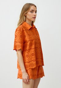 Korte mouw oranje blouse met gekartelde randen, eyelit uitsparingen en knopen aan de voorkant. Gecombineerd met bijpassende shorts met vergelijkbare details.