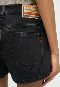 Μαύρο σορτς από τζιν με πίσω τσέπη και δερμάτινο πατς με την επωνυμία Diesel σε ταμπά χρώμα πάνω από την τσέπη στη λεκάνη ενός ατόμου.