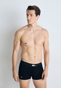 Zwarte boxershorts gemaakt van glad stof, met een brede tailleband met wit logo en een aangesloten silhouet. Het model is bovenlangs bloot.