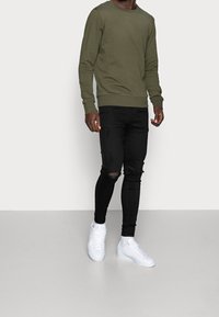 Khaki crewneck-sweatshirt, nauwsluitende zwarte jeans met gescheurde knieën, witte sneakers, gladde textuur, casual ontwerp, vooraanzicht.