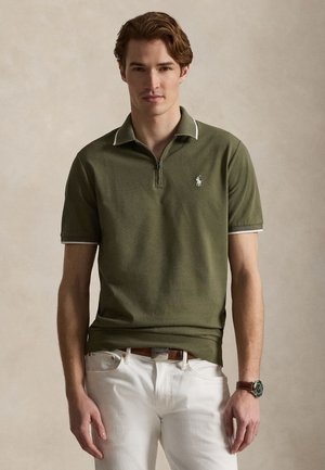 Mladić smeđe kose koji nosi maslinasto zelenu polo majicu s kratkim rukavima i bijele hlače, stoji na neutralnoj pozadini.