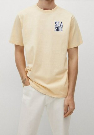 Personne portant un t-shirt beige clair avec le texte "SEA SIDE" sur la poitrine gauche et un pantalon blanc, debout avec une main dans la poche.