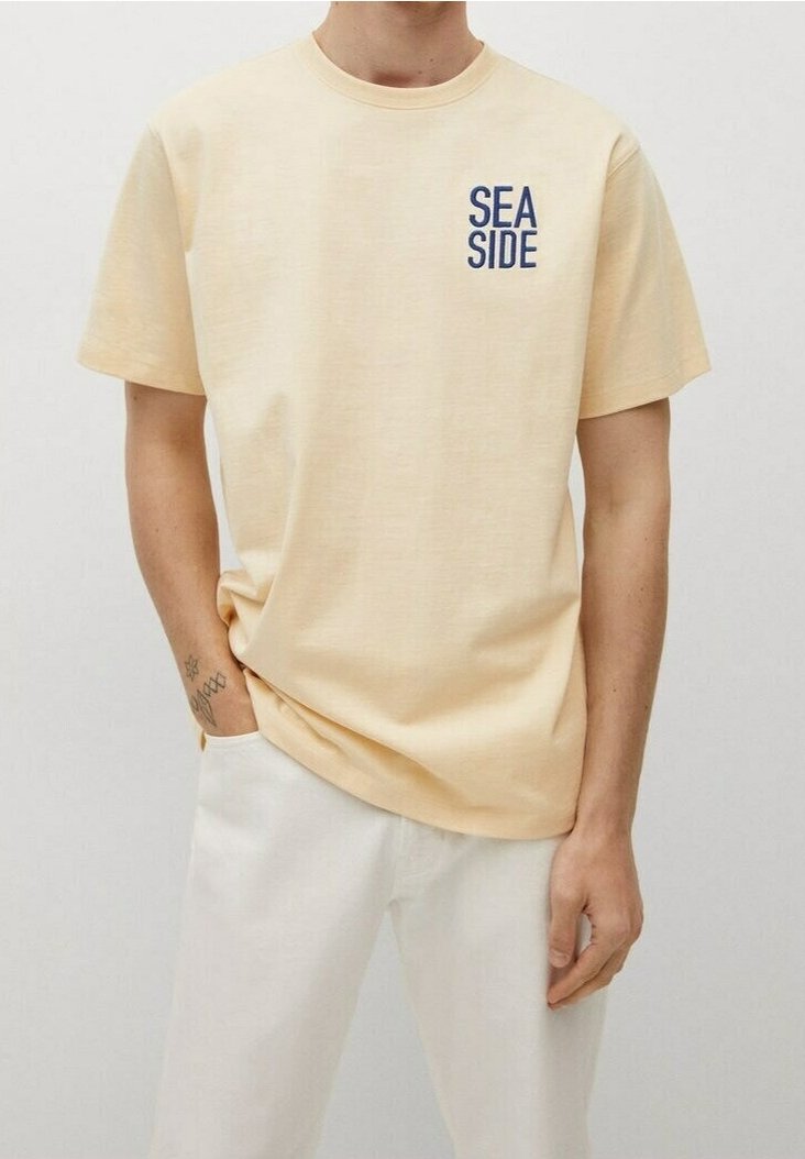 Personne portant un t-shirt beige clair avec le texte "SEA SIDE" sur la poitrine gauche et un pantalon blanc, debout avec une main dans la poche.