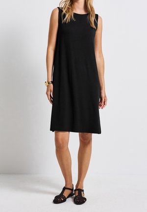 Schwarzes ärmelloses Kleid mit lockerem Schnitt, rundem Ausschnitt und knielangem Saum. Kombiniert mit schwarzen, gewebten Ballerinas. Glatte Stoffstruktur.