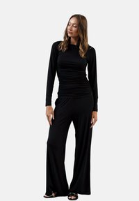Czarny kombinezon z długim rękawem z marszczonym wykończeniem w okolicy talii i szerokimi nogawkami. Gładki materiał i luźny krój. Noszony z czarnymi sandałami.