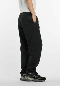 Homme portant un pantalon de survêtement noir et des baskets noir et gris, se tenant avec une main dans la poche contre un fond blanc uni.