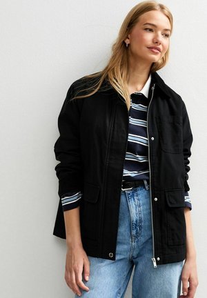 Chaqueta negra con cremallera frontal, dos bolsillos delanteros y un cuello. Llevada sobre una camisa a rayas azul y blanca y unos pantalones vaqueros de cintura alta azul.