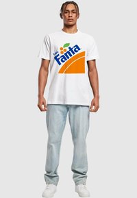 Hvid bomulds t-shirt med et stort blåt og orange "Fanta" logo. Model iført lyseblå jeans og hvide sneakers. Enkel, afslappet design.