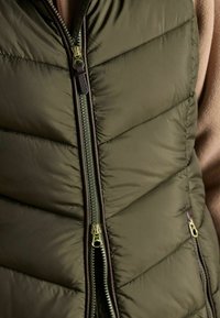 Oliwkowa kamizelka puchowa wykonana z lekkiego materiału, z pełnym zamkiem błyskawicznym z złotym wykończeniem oraz teksturą w ukośny wzór.