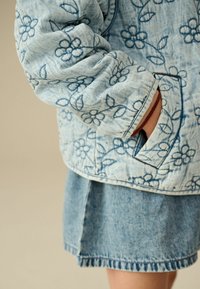 Hellblaues Denim-Jacket mit floraler Stickerei, ausgestattet mit einer gesteppten Textur und gerippten Bündchen. Kombiniert mit einem passenden Denim-Rock.