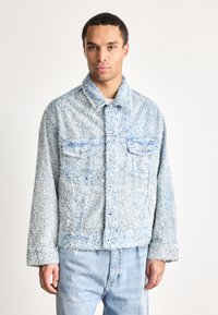 Man wearing a light blue textured button-up jacket with two chest pockets and light blue jeans, standing against a plain background.