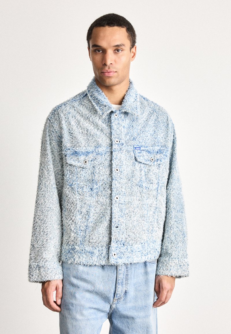 Man wearing a light blue textured button-up jacket with two chest pockets and light blue jeans, standing against a plain background.