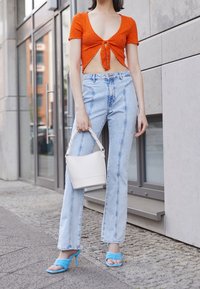 Pomarańczowy prążkowany krótki top z wiązaniem z przodu, jasnoniebieskie dżinsy z wysokim stanem, niebieskie sandały na obcasie oraz biała torebka o fakturze.