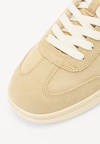 Beige suède sneaker met een ronde neus, crème veters en complexe stiksels langs de zool. Gladde textuur en subtiele ontwerpelementen.