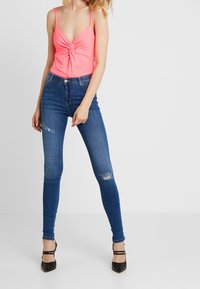 Neon růžový top bez rukávů s uzlovým detailem v přední části, spárovaný s tmavě modrými skinny džíny s trhlinami na stehnech a černými podpatky.