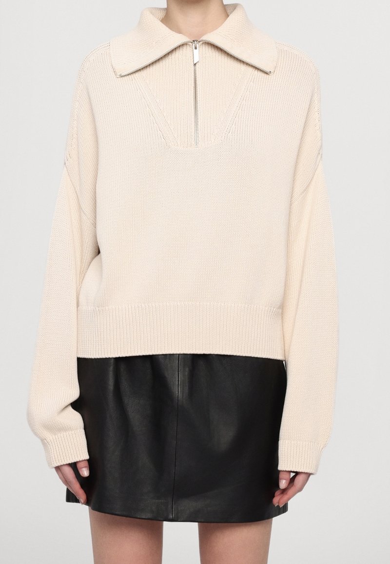 Pull tricot crème avec col à fermeture éclair au quart, associé à une mini-jupe en cuir noire portée par une personne debout sur un fond uni.