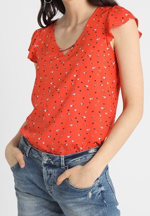 Kvinde iført en orange kortærmet bluse med små hjerte-mønstre og blå jeans med hænderne i lommerne.