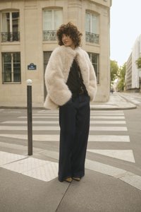 Šviesiai smėlinis dirbtinės kailio paltas, dėvimas ant juodo bluzono su nėriniais, derinamas su plačiomis tamsiai džinsinėmis kelnėmis ir smėliniais aukštakulniais, stovinti prie perėjos.