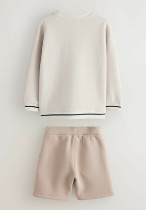 Camiseta de punto waffle de manga larga beige con puños a rayas negras, combinada con pantalones cortos de cintura elástica en beige claro, sobre fondo blanco.