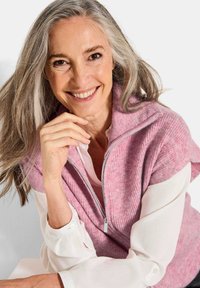 Rosa gestrickte Weste mit Reißverschluss, bestehend aus einer gerippten Textur und einem umgeschlagenen Kragen, geschichtet über einem weißen Hemd mit Knopfmanschetten.