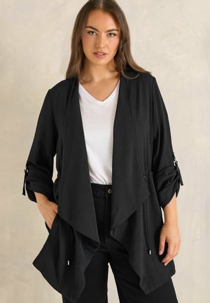 Mujer con chaqueta negra de frente abierta con las mangas remangadas, sobre camiseta blanca de escote en V y pantalones negros, de pie contra un fondo liso.