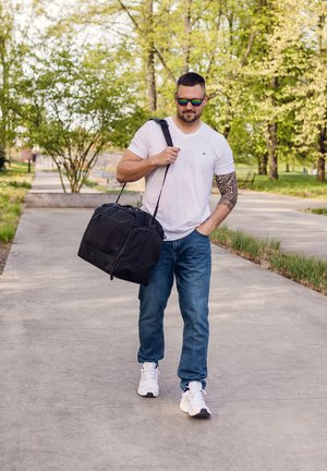 Black duffel bag with a top zipper and shoulder strap, worn by a person in a white t-shirt and blue jeans, outdoors on a walkway.
