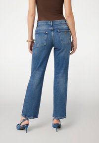 Aukšto liemens mėlyni džinsai su tiesiomis kojomis. Yra dvi užpakalinės kišenės ir mažas logotipo etiketė. Dėvimi su mėlynomis aukštakulnėmis basutėmis.