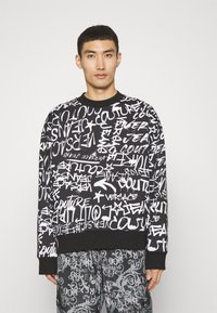 Felpa nera con testo e motivi in stile graffiti bianchi. Collo rotondo e polsini a coste. Indossata con pantaloni grigi a fantasia.