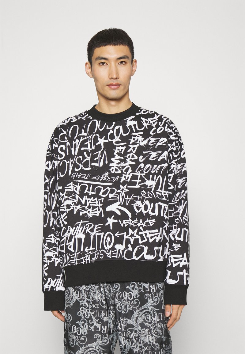 Felpa nera con testo e motivi in stile graffiti bianchi. Collo rotondo e polsini a coste. Indossata con pantaloni grigi a fantasia.