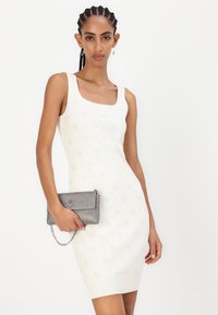 Pletené bodycon šaty v krémové barvě s diamantovým vzorem, s čtvercovým výstřihem. Modelka drží malou stříbrnou kabelku s řetízkovým detailem.
